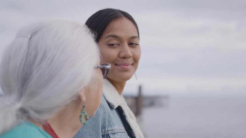 A younger woman with dark hair turns her head and smiles warmly at an older woman with gray hair tied in a ponytail as they both stand facing a blurred waterfront in the background