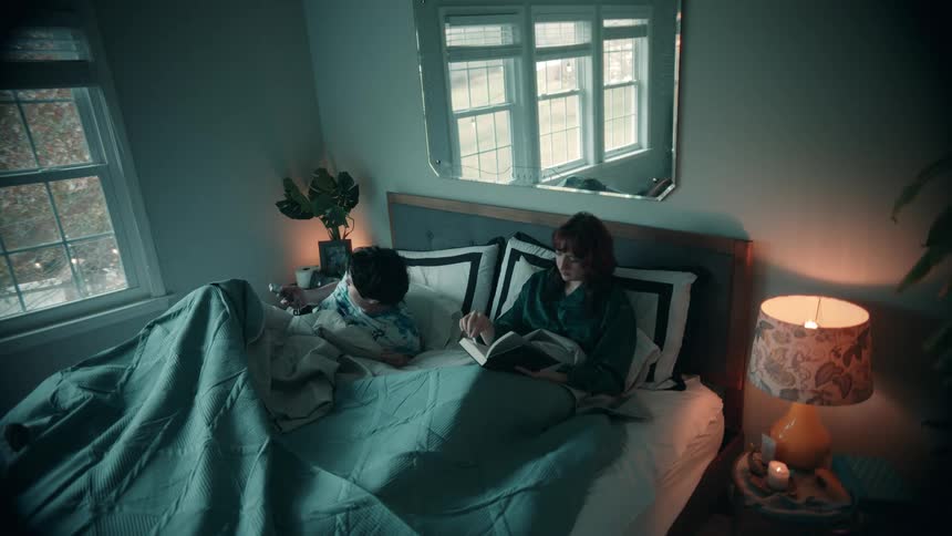 A wide shot shows a couple lying in bed in a dim room the woman sits up with a book and the man lies under the covers briefly looking at his mobile phone before turning to rest