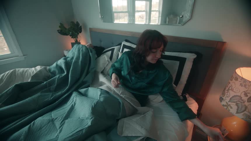 A woman sitting up in a large bed next to a sleeping man looks down at her hands before reaching over to a nearby glowing lamp and then she picks up a book and starts reading quietly while still covered by the bedding