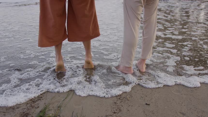Two women stand barefoot on the beach as waves roll in over their feet