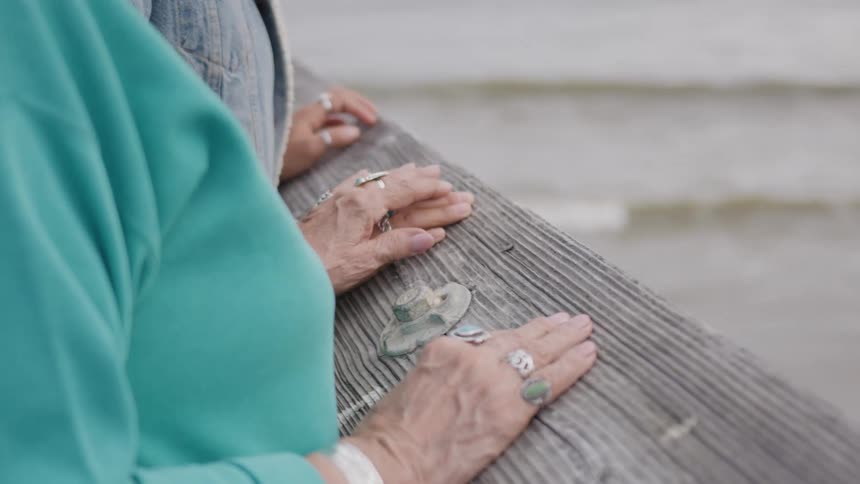 Two pairs of hands holding hands rest on a weathered wooden railing overlooking a blurred area