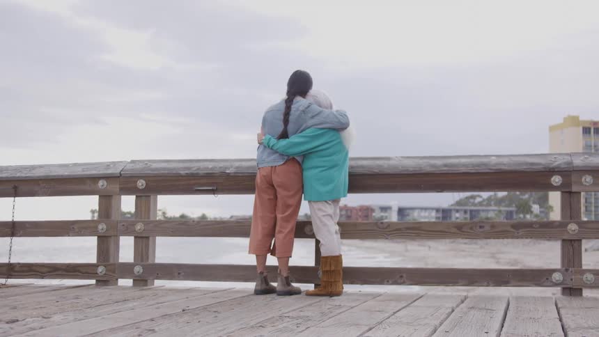 A rearview shot of two women one younger and one older standing on a wooden deck overlooking a beach and distant buildings as they tightly embrace each other