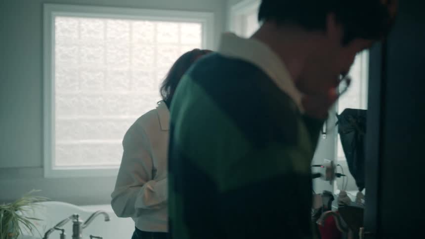 A man and a woman in a brightly lit bathroom laugh together as the woman gently touches the mans cheek then they turn away from the camera toward the mirror and continue their conversation