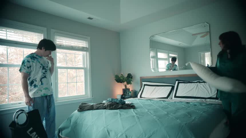A woman wearing dark green pajamas makes the bed by adjusting pillows on the right side while a young man in casual sleepwear picks up a laundry bag in a brightly lit modern bedroom