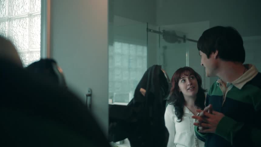 A man wearing a green and black striped rugby shirt and a woman with red hair and a white shirt are standing in the foreground looking toward the left the man is smiling and adjusting his hair on the left side mostly obscured and out of focus is anot