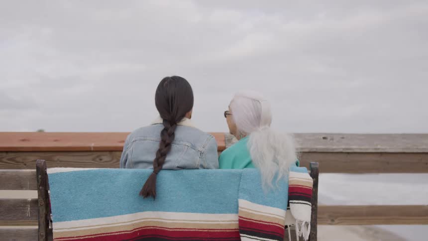 Two people sit on a seaside bench chatting while looking at the horizon
