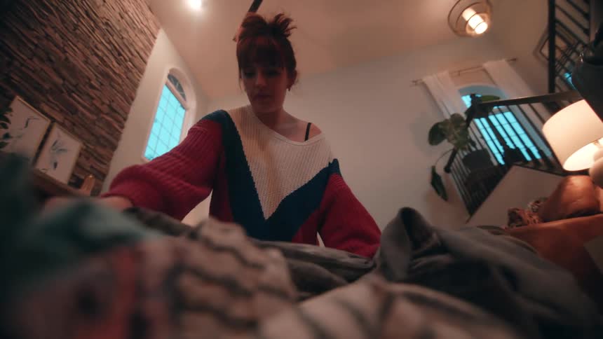 A woman with her hair in a bun wearing a colorful sweater sitting on the floor and focused on folding a large pile of clothes in a home interior with a textured brick accent wall and a nearby staircase