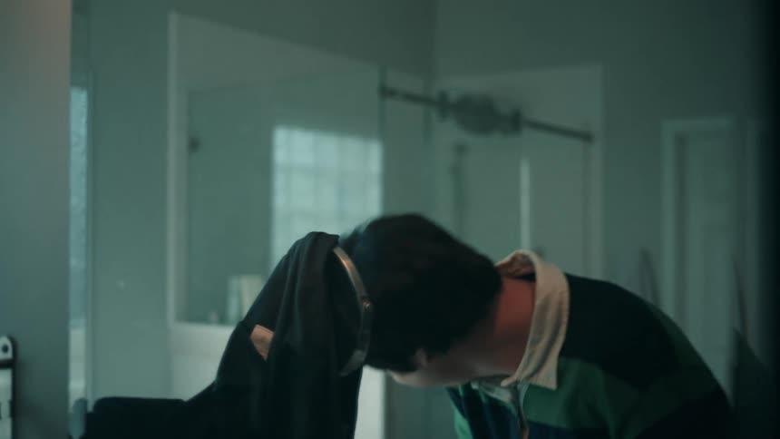 A young man laughs as he rubs the front of his hair looking in a mirror in a bathroom