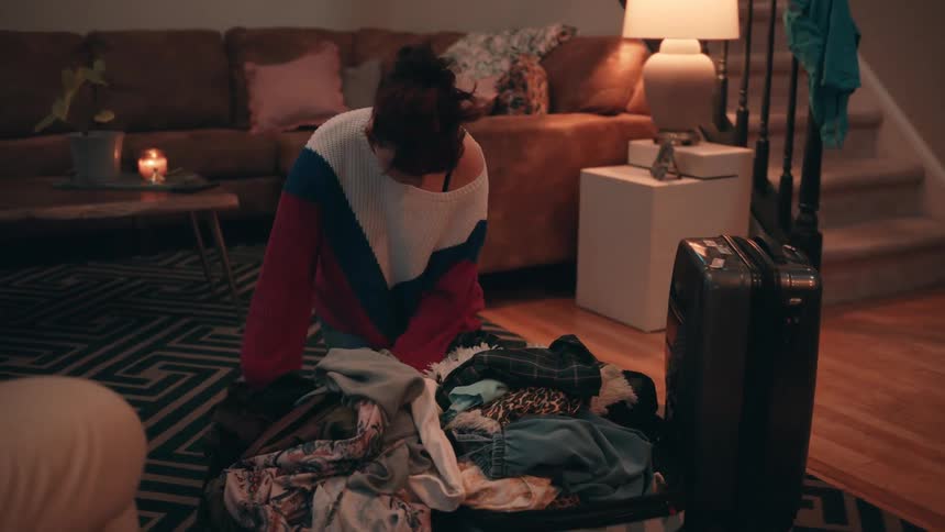 A woman in a red white and blue sweater is kneeling on a rug packing a black suitcase surrounded by a large pile of clothes in a cozy living room