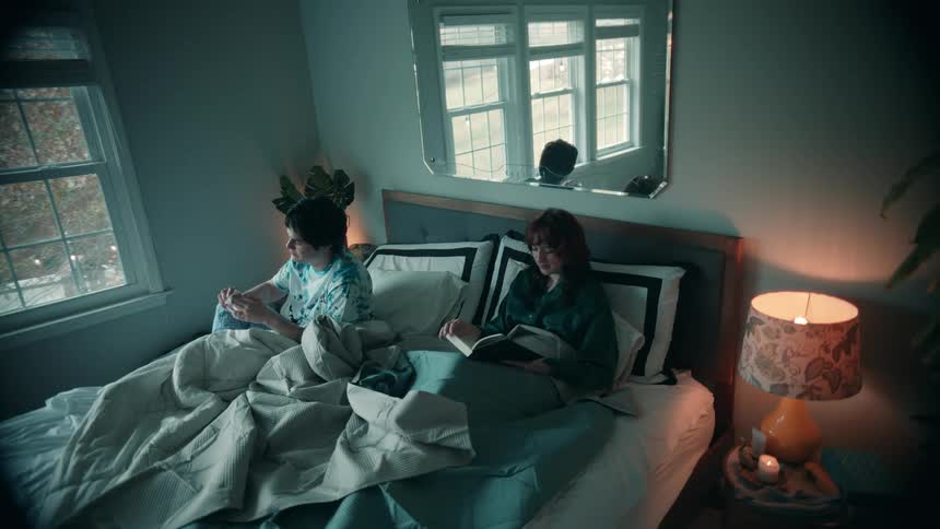 Man sneezes and gets out of bed while another woman reads a book beside him
