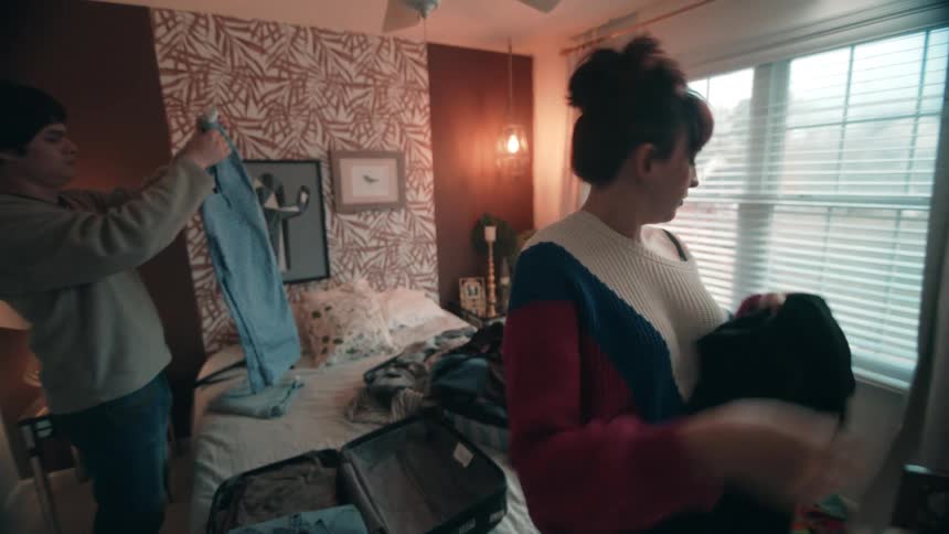 A man and a woman are in a cozy bedroom with patterned wallpaper and a window with blinds standing on opposite sides of a bed covered in clothes as the man holds up a pair of pants and the woman sorts through a pile of clothing items