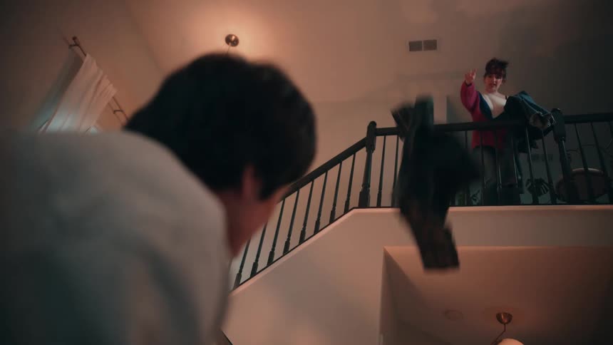 Woman on the upper floor throws clothes while another watches from the bottom of the stairs