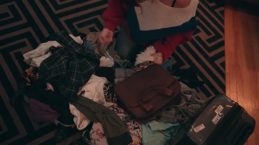 A highangle shot looks down upon a woman wearing an offtheshoulder sweater as she kneels on a geometric patterned rug actively digging through a large messy pile of clothing