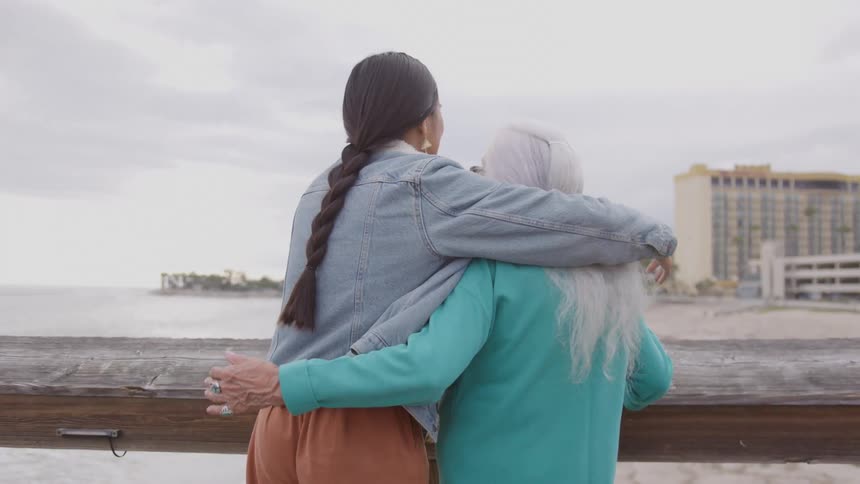 Two people with arms around each other look out over the coastal view from a deck
