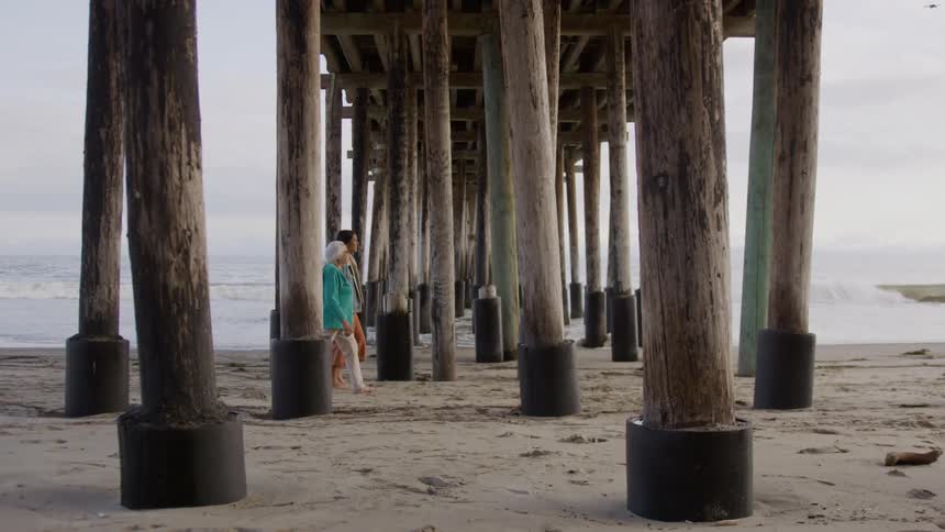 Couple walks beneath a pier structure on the sand near the sea