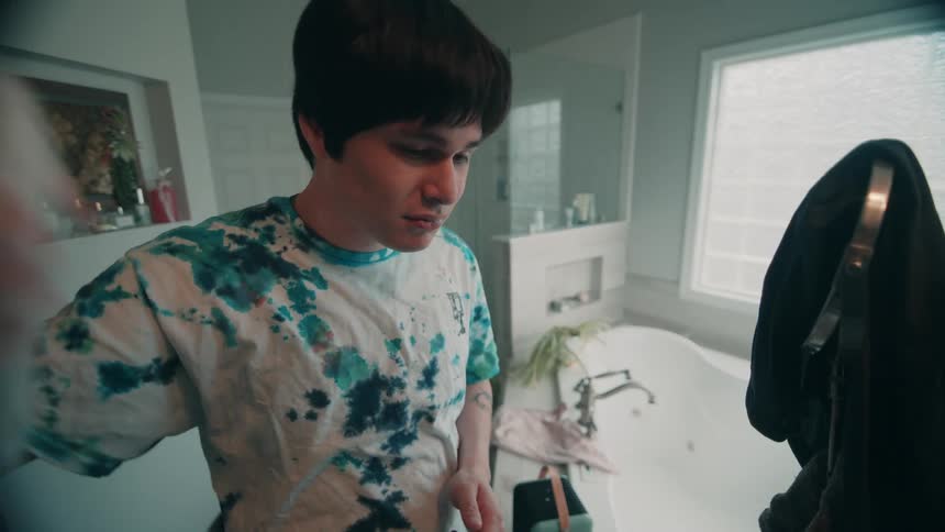 The video shows a person with dark hair wearing a white tshirt with blue and green tiedye patterns standing in a bathroom they are brushing their hair with a red brush and then use a spray bottle on their haira bathtub and a large window are visi