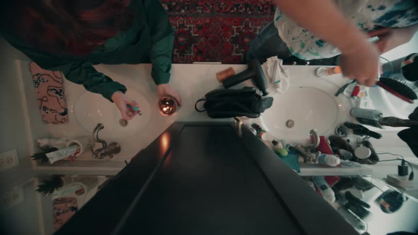 Shot from an unusual overhead angle two individuals stand at a double bathroom sink countertop engaging in a shared morning routine using various toiletries and eventually lighting a small candle
