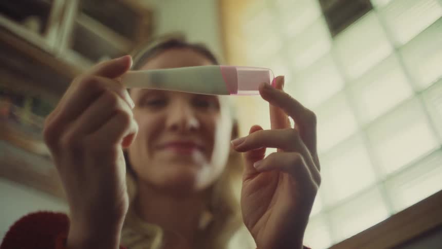 A womans hands as she holds up a white and pink pregnancy test with the device and her hands sharply in focus and her face blurred in the background bathroom