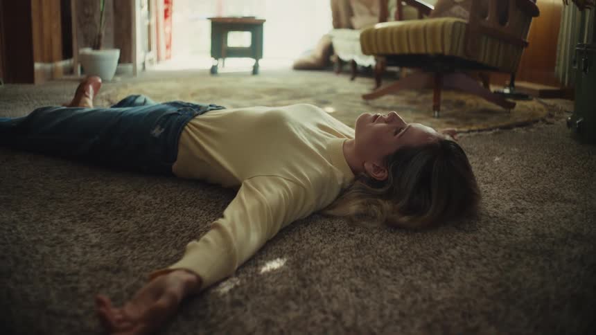 A caucasian female with light hair lies face up on a carpeted floor in a living room with furniture visible in the background
