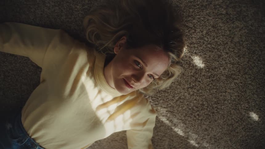 A woman with blonde hair is looking forward with a gentle content expression while lying on a brown carpet