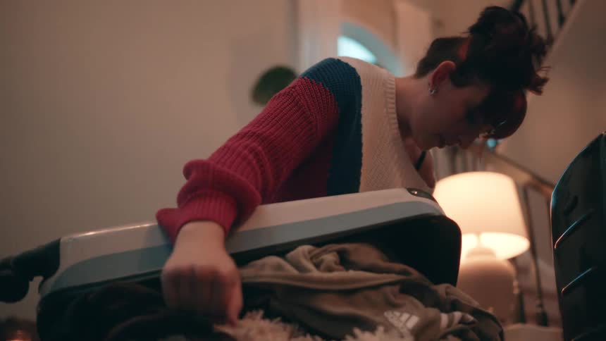 A young woman in a colorful knit sweater kneels over a suitcase overflowing with clothes struggling to pack in a dimly lit home entryway
