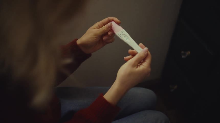 A caucasian persons hands holding a positive pregnancy test with the person looking forward against a dark indoor background