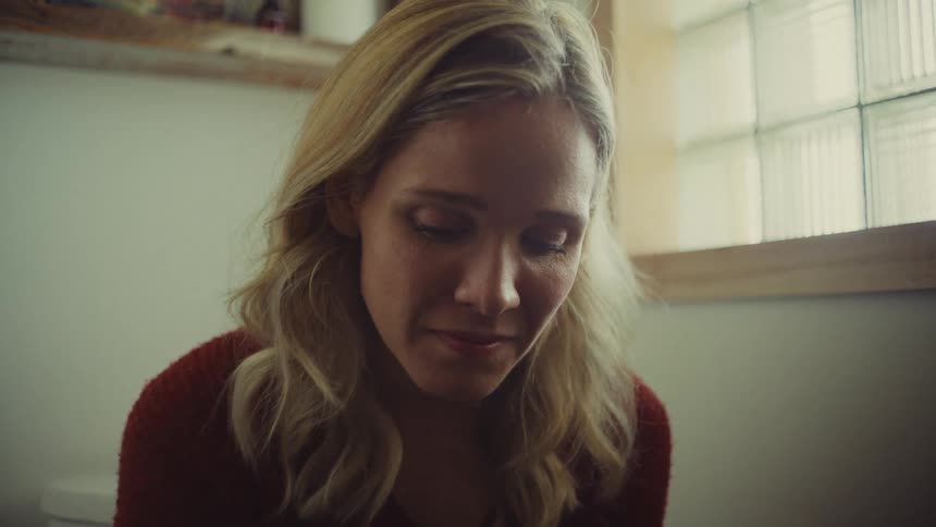 Young woman wearing a red sweater seating on a toilet with a sad and nervous facial expression indoors during day time