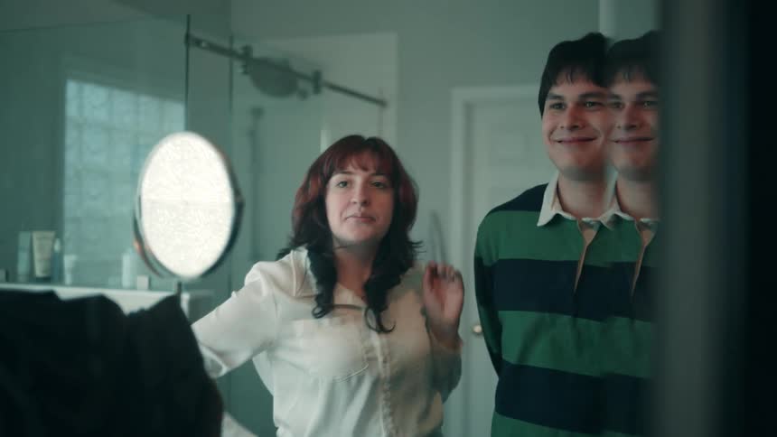A woman helps a man adjust the collar of his shirt in a well lit modern bathroom with their reflection visible in the mirror