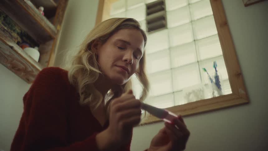 A blonde woman in a red sweater looks down intently at a pink and white pregnancy test stick held in her hands near a window