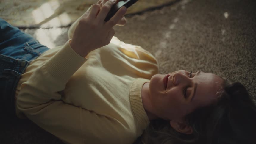 Young woman lying down on the floor texting on a smartphone and smiling indoors during day time