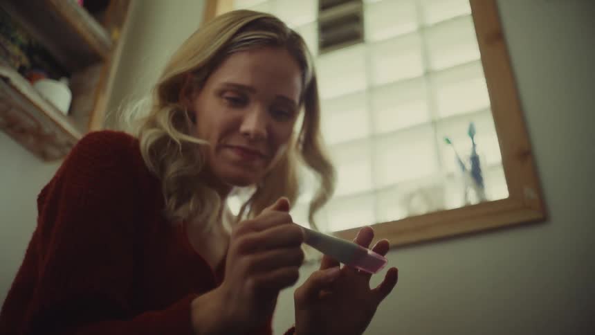 A blonde woman in a red sweater is captured in a lowangle closeup shot as she intently examines a pregnancy test in her hands and has a happy reaction to the test result in a dimly lit bathroom