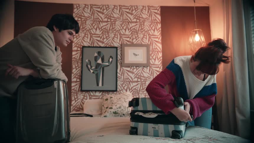 Two people try to close overfilled suitcases placed on the bed