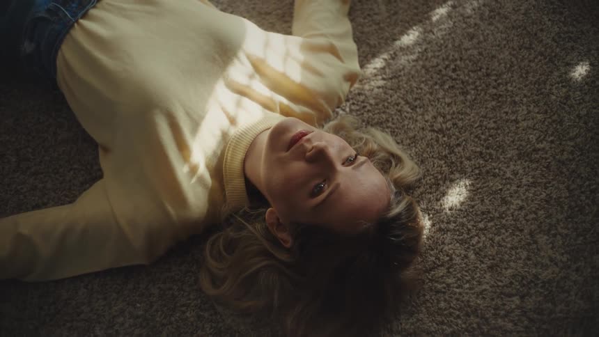 This video shows a woman lying on her back on a brown carpet wearing a light yellow sweater and blue jeansrays of sunlight stream in creating stripes of light across her body and the floor as she opens and closes her eyes while looking up at the cam