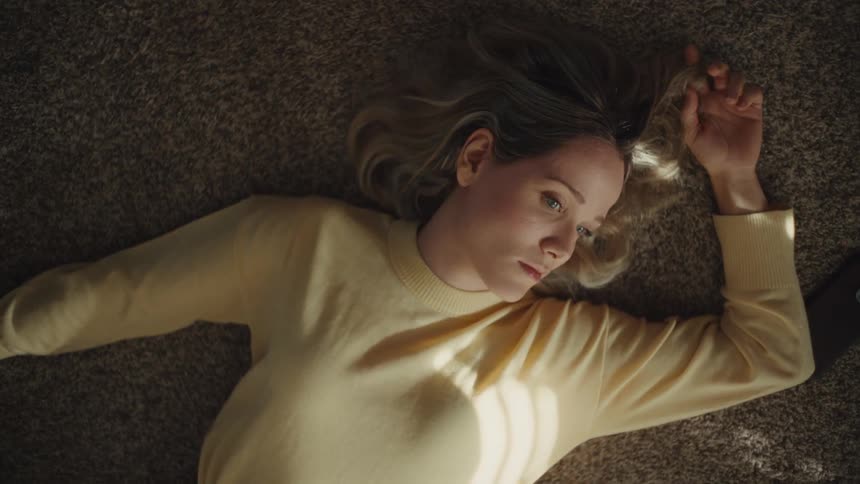 A woman with blonde hair lying on a textured brown carpet in a yellow sweater and jeans resting her arm above her head as she looks upward with a thoughtful expression while illuminated by a diagonal beam of sunlight