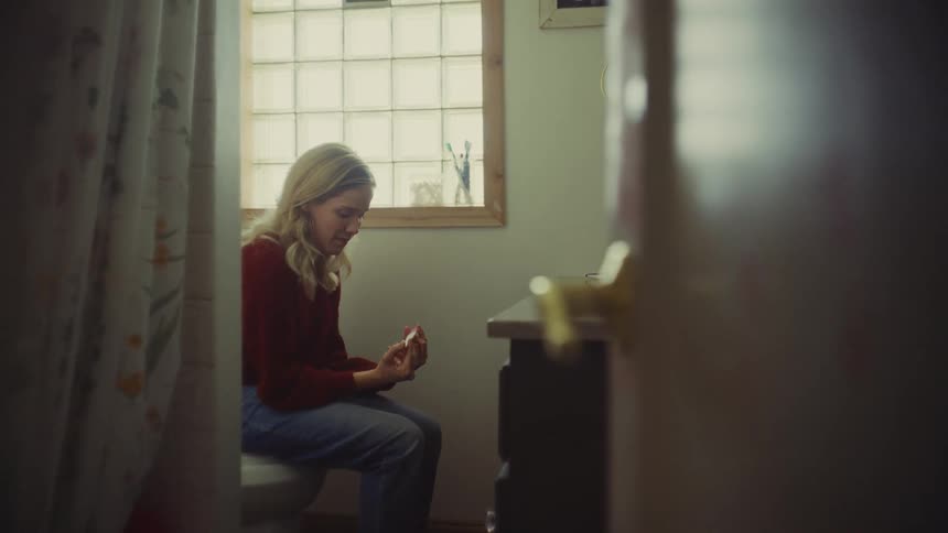 A young blonde woman wearing a red shirt and jeans sits on a toilet in a brightly lit bathroom holding and intently staring at a pregnancy test in her hands with an anxious expression framed from the side through a partially open doorway