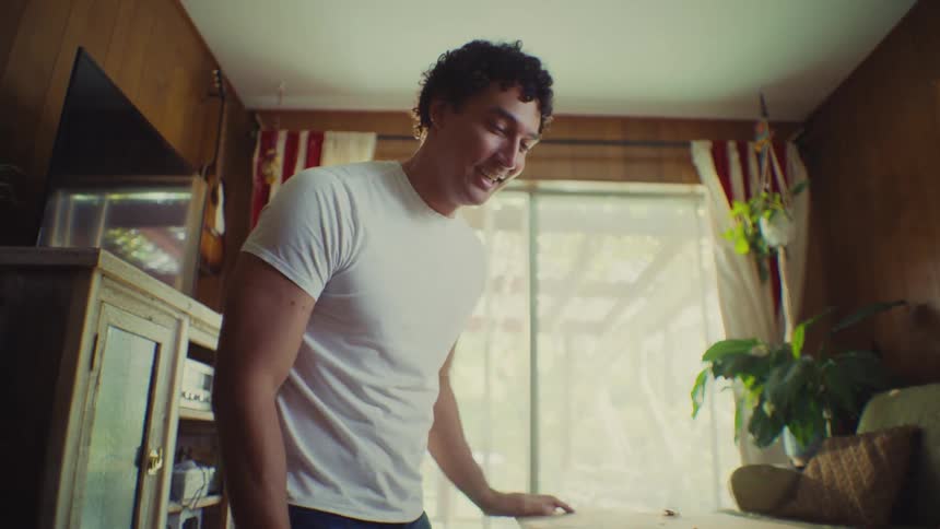 This short clip shows a young man in a casual white tshirt reacting with excitement and joy as he opens his hands above a table the background suggests a cozy slightly vintage living space with wood paneling and striped curtains