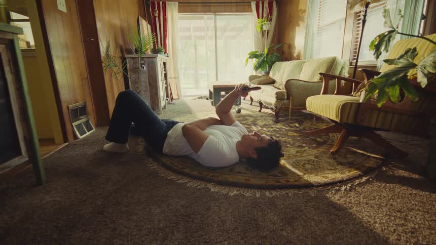 Young man wearing a white shirt and jeans lying down on the floor of a living room using a smartphone indoors during day time