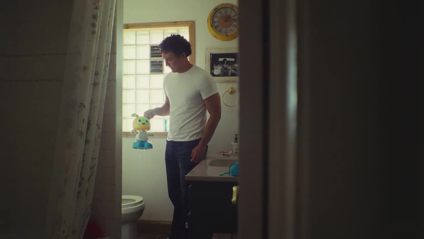 A man in a white tee shirt and jeans is standing in a brightly lit bathroom holding a colorful toy he picked up from inside the toilet
