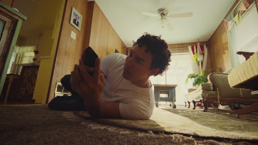 A man with curly hair wearing a white shirt is seen lying on his back on a patterned rug in a woodpaneled room actively engaging with his smartphone before sitting up quickly and reacting with a sudden surprised movement