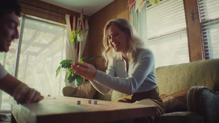 This video shows two people sitting across a low wooden table in a retrostyle living room playing a game involving dicea woman with light hair wearing a light blue top and brown pants rolls the dice and then reacts with an exaggerated expression of e
