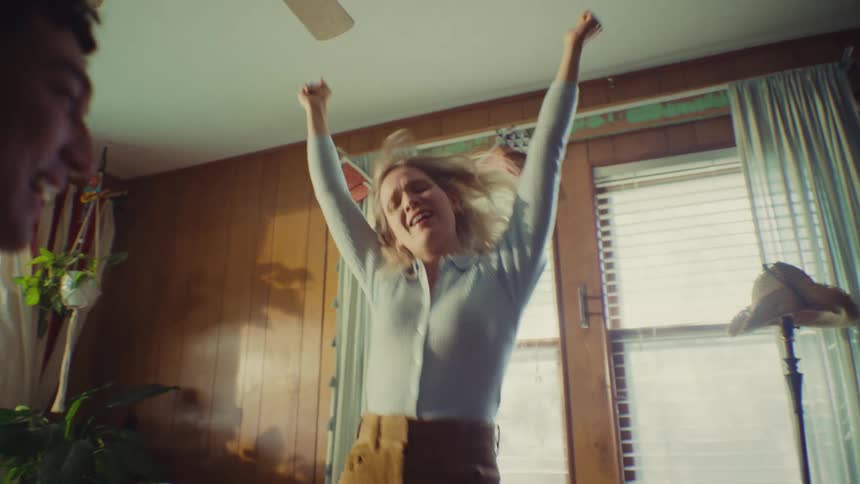 A young couple enjoys their time in the living room the woman jumps excitedly on the couch while the man tosses pieces of paper into the air