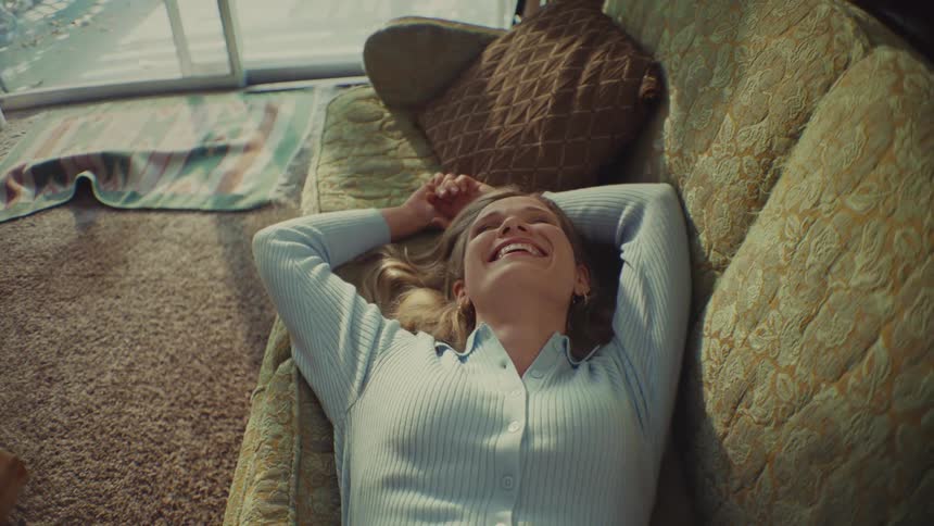 A smiling woman with curly hair wearing a light blue ribbed sweater is lying comfortably on an olive green patterned couch in a sunlit room