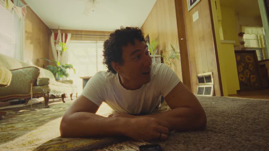 A young man with curly hair and a white shirt lies on a rug and interacts with a phone on the floor in a retro style sunlit living room