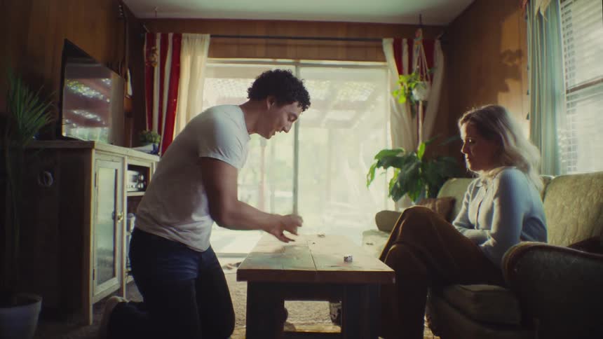 A man and a woman are playing a game on a coffee table in a rustic living room with the woman celebrating after the man collapses in mock defeat