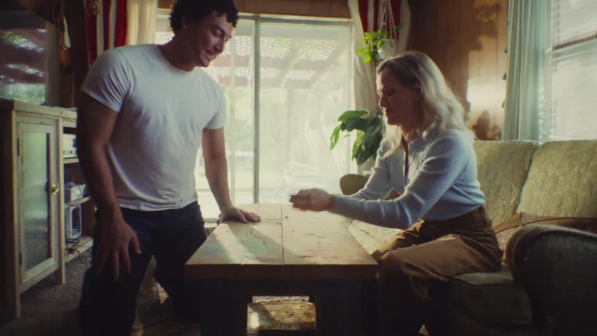 A man and a woman are enthusiastically playing a board game or tabletop game in a living room filled with natural light the woman covers her face and then laughs in excitement as the man shakes dice indicating a fun and competitive game moment
