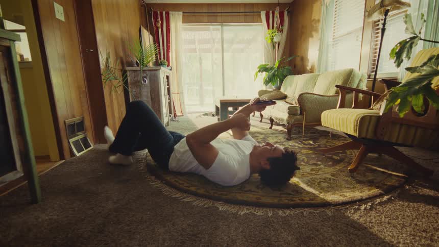 A man wearing a white shirt and dark pants is lying on his back on the floor resting his head on a small rug while looking up and holding a smartphone in a retrostyled living room