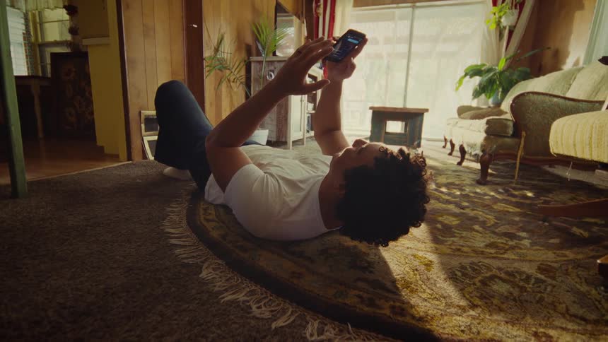 A man is laying on his back on a rug in the living room yelling at his phone