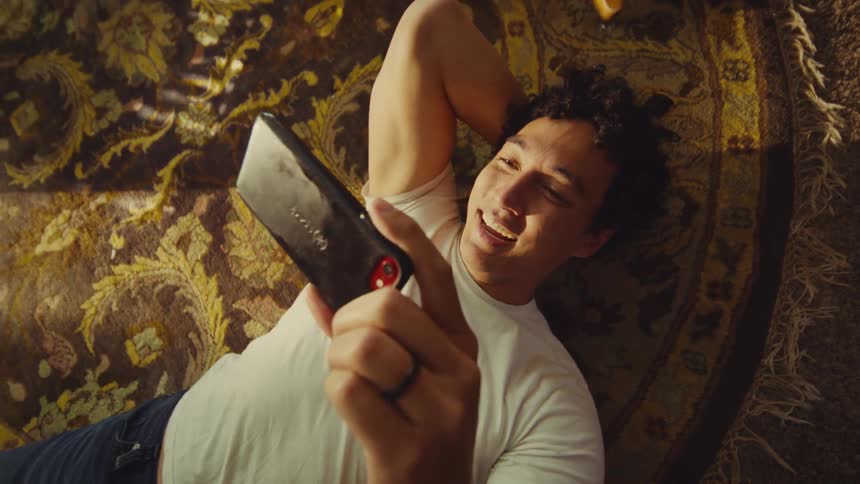 A very happy man cheering in a white tshirt is looking at a smart phone in his hand while lying on a patterned rug in warm light