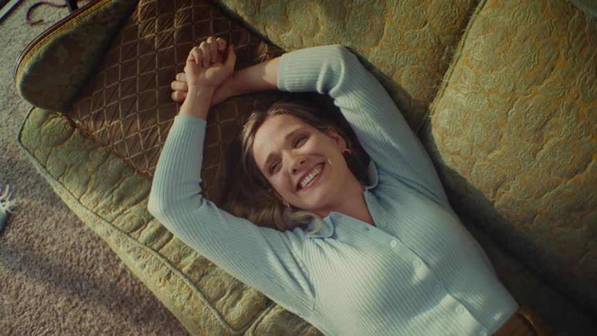 A blonde woman wearing a light blue buttonup cardigan lies relaxed on a patterned olive green sofa stretching her arms above her head while looking up and smiling warmly toward the camera