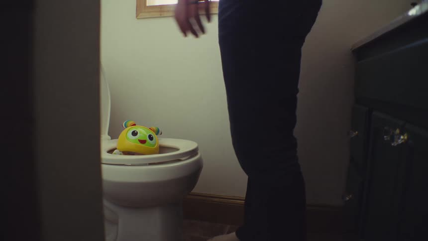 A man stands next to a toilet with the seat up looking down and picking up a brightly colored yellow and green childs toy resting inside the bowl